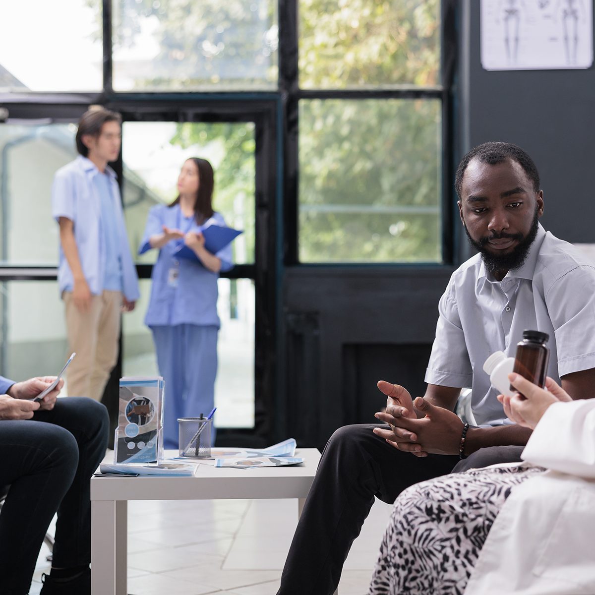 Elderly medic showing drug bottles to patient discussing health care treatment during medical consultation in hospital waiting area. Multi ethnic people standing in lobby before start examination